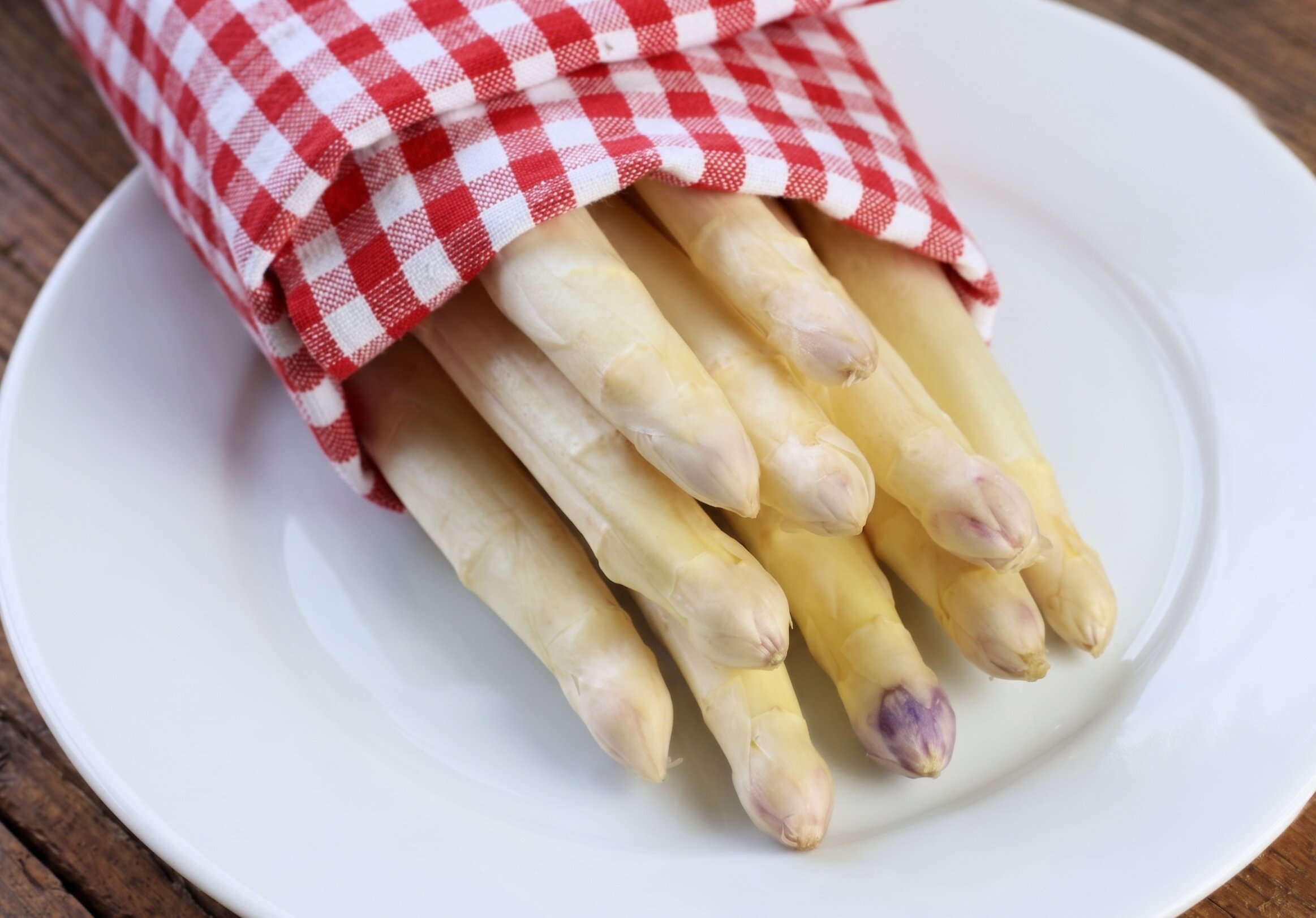 Eine Nahaufnahme eines Bündels frischer weißer Spargelstangen, die auf einem sauberen weißen Keramikteller liegen. Der Spargel ist teilweise in ein rot weiß kariertes Gingham Tuch gewickelt. Die Stangen sind cremeweiß und haben fest geschlossene, helle Spitzen.